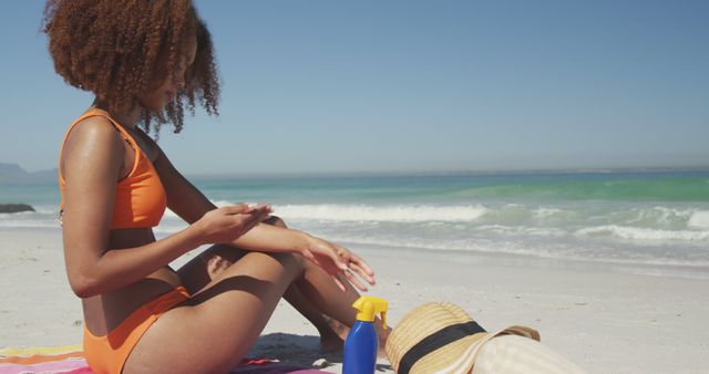 Young woman in orange swimwear applying sunscreen on a sunny beach with ocean background. Ideal for content related to summer vacations, beach lifestyle, skincare routines, health and wellness tips.