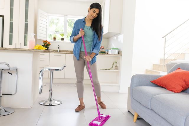 Young woman mopping floor in modern living room, showcasing daily housework and hygiene. Ideal for use in articles or advertisements related to cleaning products, home maintenance, and lifestyle blogs focusing on domestic life and routines.