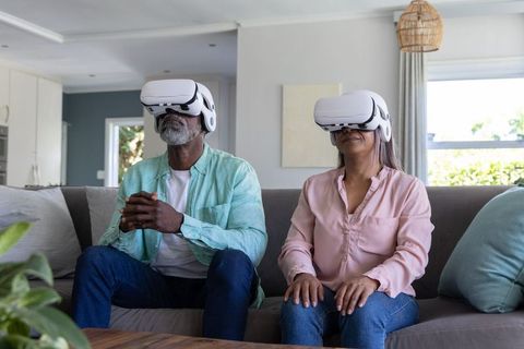 Diverse senior couple experiencing virtual reality at home with white VR headsets on sofa