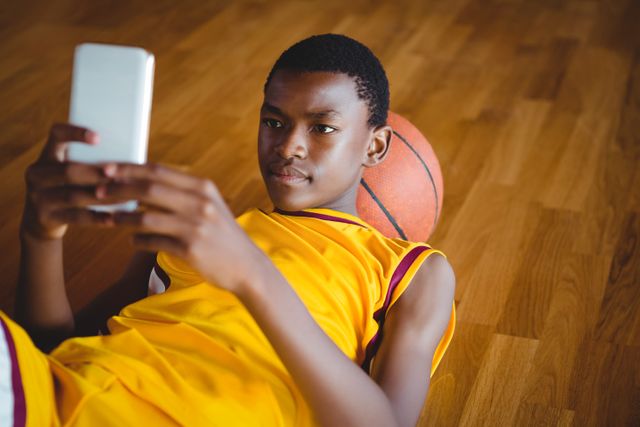Teenage boy in a yellow sports jersey lying on a hardwood basketball court, using a mobile phone. Ideal for themes related to youth, technology, sports, relaxation, and leisure activities. Suitable for articles, advertisements, and social media posts focusing on teenage lifestyle, digital engagement, and sports culture.