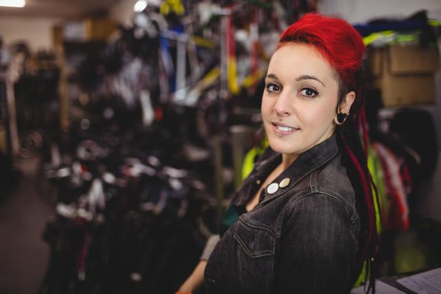 Young female mechanic with red hair smiling confidently in a workshop. She is wearing casual clothing and has an alternative style with piercings and tattoos. Ideal for use in articles about women in trades, alternative fashion, and workplace diversity.