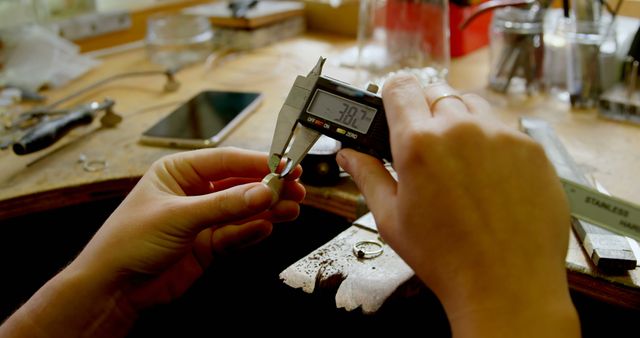 Close-up of a craftsman using a digital caliper to measure a piece of jewelry in a workshop. Useful for illustrating craftsmanship, precision work, and artisanal processes in articles, instructional materials, or blogs about jewelry making, metalworking, and fine craftsmanship.