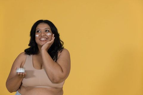 Plus-size woman applying moisturizer smiling and touching cheeks on mustard yellow backdrop