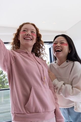 Diverse female friends wearing pink gel eye patches laughing and posing in sunlit modern apartment