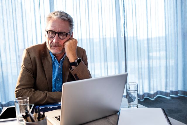 Senior businessman sitting at office desk, looking sad and thoughtful. He is wearing glasses and a brown blazer, with a laptop and a glass of water on the desk. This image can be used to depict workplace stress, corporate challenges, or the emotional side of business life. Suitable for articles on mental health, business environments, or professional stress management.