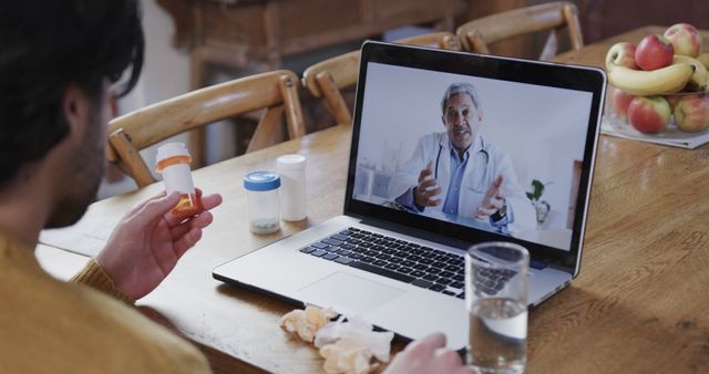 Caucasian man with pills on table having laptop video call with biracial male doctor at home. Domestic life, medicine and communication, unaltered.