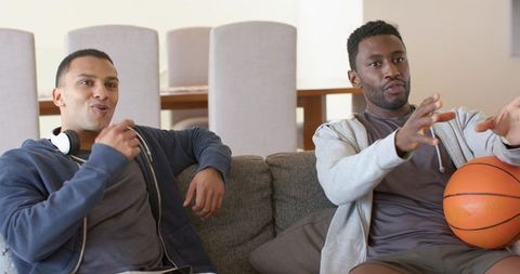 Two male friends watching game on sofa holding basketball cheering and talking casually
