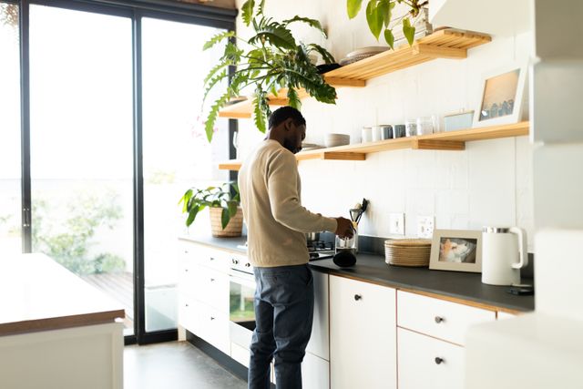 African American man pouring coffee in a modern kitchen with wooden shelves and plants. Ideal for use in lifestyle blogs, home decor websites, and advertisements promoting inclusivity and modern living.