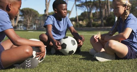 Diverse youth soccer teammates sitting on field holding ball during sunny park practice