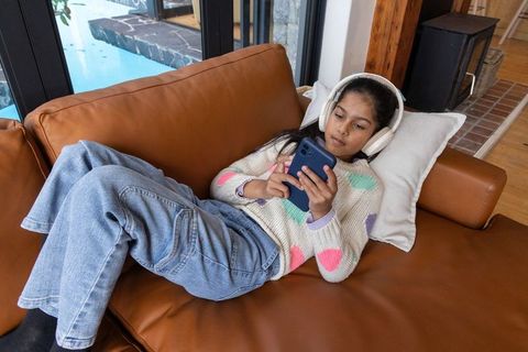 Indian teenage girl relaxing on leather sofa using smartphone and headphones by pool