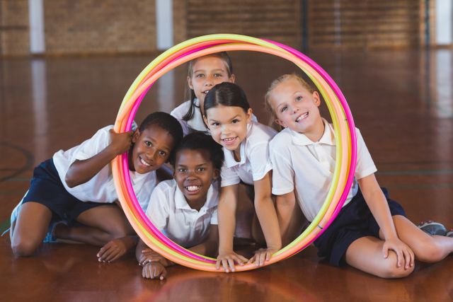 Diverse group of school kids smiling and looking through a colorful hula hoop in a gym. They are wearing school uniforms and appear happy and energetic. Ideal for use in educational materials, advertisements for school programs, or articles about childhood fitness and teamwork.