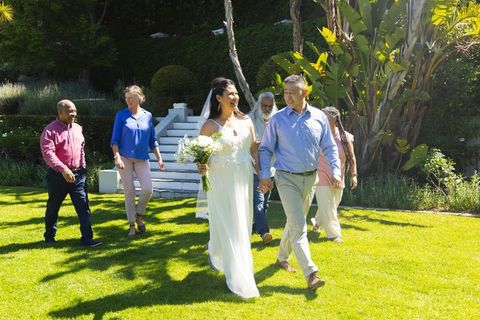 Asian bride and groom walking with family in sunlit garden wedding celebration