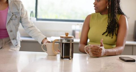 Diverse women chatting and sipping coffee at kitchen counter with french press