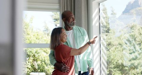 Mature couple admiring mountain view through sunlit glass door embracing future