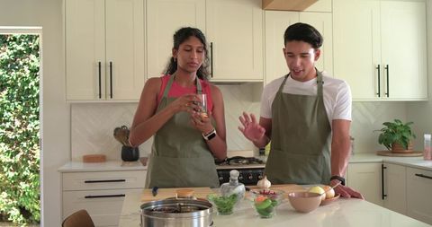 African American couple cooking together in sunlit modern kitchen wearing olive aprons