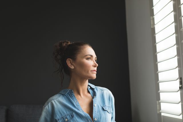Woman standing indoors, gazing outside through window shutters, wearing a denim shirt. Natural light illuminates her face, creating a serene and contemplative atmosphere. Ideal for use in lifestyle blogs, mental health articles, home decor advertisements, and personal reflection themes.