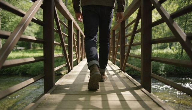 Walking man crossing sunlit wooden footbridge over stream in lush forest outdoors
