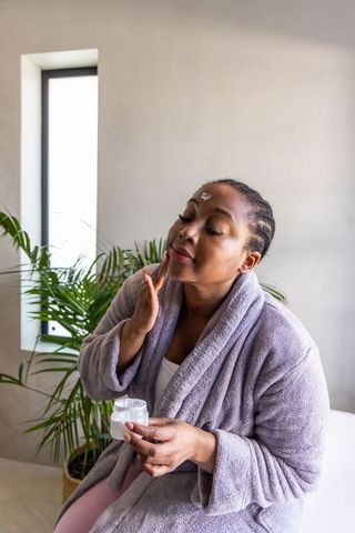 Middle-aged african american woman applying facial cream in bathrobe near sunlit window