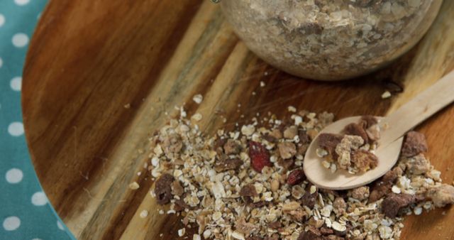 Close-up of muesli spilled on wooden table next to glass jar and wooden spoon, emphasizing texture and diversity of ingredients. Suitable for use in culinary blogs, nutrition guides, healthy eating promotions, recipe books, food articles, and breakfast product advertisements.