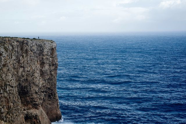 A lone figure is standing on the edge of a high coastal cliff, looking out at the expansive ocean under a partly cloudy sky. The sea appears slightly turbulent with visible waves. This image is ideal for concepts of solitude, contemplation, travel, adventure, freedom, and the beauty of nature. Perfect for use in travel magazines, inspirational marketing, blogs, or social media posts highlighting scenic landscapes and solitary experiences.