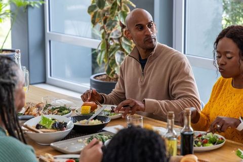 Diverse family enjoying casual daylight lunch around table, man serving salad and corn