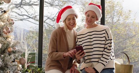 Mother and daughter wearing santa hats sharing phone by frosted christmas tree - cozy holiday moment