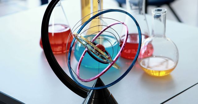 A gyroscope sits on a table surrounded by colorful liquid-filled flasks in a science laboratory setting, with copy space. Gyroscopes are used to demonstrate principles of angular momentum and stability in physics.