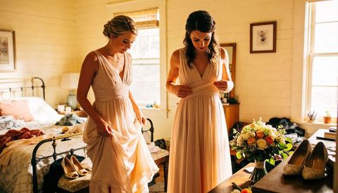 Bridesmaids adjusting peach pleated gowns in sunlit rustic bedroom preparing for wedding