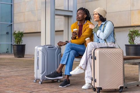 Young couple waiting with carry-on suitcases and coffee outside modern transport terminal