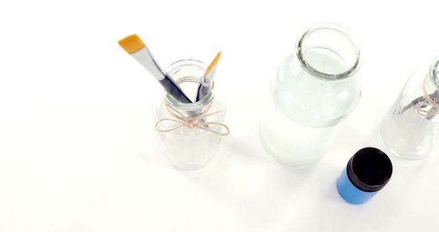 Clear glass jars with water and paintbrushes tied with twine on white surface, perfect for illustrating artistic hobbies, creative workspaces, craft tutorials, and DIY projects.