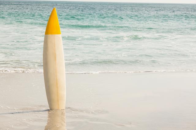 Surfboard standing upright on sandy beach with gentle ocean waves in background. Ideal for use in travel brochures, surfing advertisements, summer vacation promotions, and beach-themed designs.