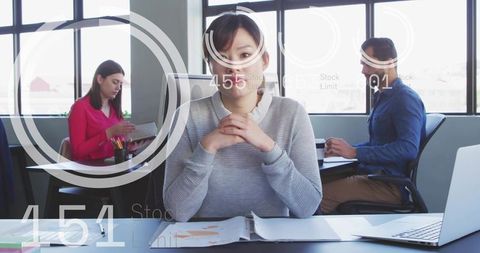 Woman sitting at desk clasping hands over documents with laptop in modern daylight office