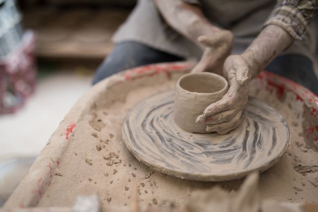 Male potter molding clay on a pottery wheel in a workshop. The image focuses on the hands and the clay, highlighting the craftsmanship and skill involved in pottery making. This image can be used for articles on traditional crafts, artistic skills, pottery classes, or handmade ceramics.