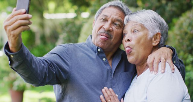 Senior couple standing outside making silly faces while taking a selfie. Perfect for campaigns promoting senior lifestyle, joyful living, self-expression, or casual family photography. This entertaining image can be used in advertisements, social media posts, or brochures for products or services targeting older adults and family activities.