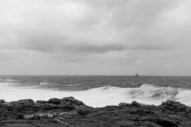 Black and white scene of a stormy sea with choppy waves crashing against a rocky shoreline. Clouds loom overhead, creating a dramatic sky. A distant ship is visible on the horizon, hinting at rough maritime conditions. This image is suitable for use in projects related to nature, marine weather, coastal landscapes, and dramatic seascapes.