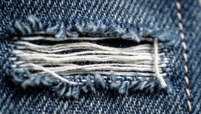 Macro showing frayed denim tear exposing white weft threads and tan stitching blue twill