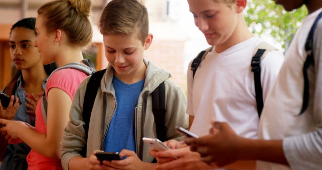 Image of a diverse group of teenagers engaging with their smartphones while standing outdoors. All are focused on their devices, indicating an emphasis on connectivity and digital communication. This image can be useful for articles or advertisements related to teenage behavior, social media influence, technology in education, or the importance of staying connected. Ideal for use in blogs, social media campaigns, educational materials, and advertisements targeting young teenagers.