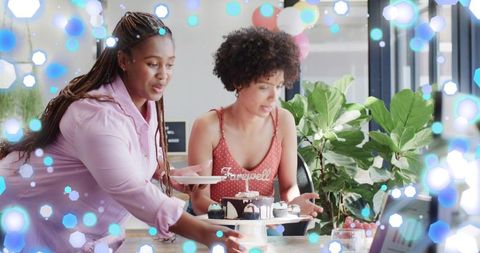 Colleagues arranging farewell cake and cupcakes on office table with laptop, plants