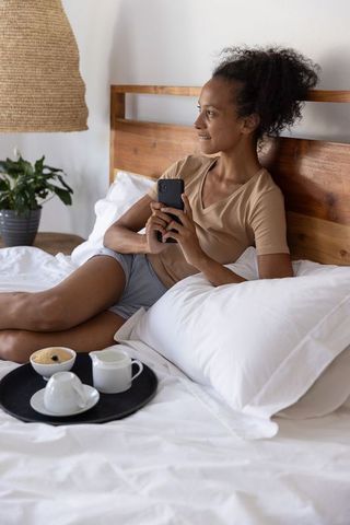 Relaxing african american woman reclining on bed holding smartphone and breakfast tray