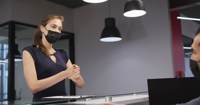 Receptionist wearing mask assisting client in modern office with safety precautions. Suitable for materials related to business operations during COVID-19, office safety, customer service, and workplace hygiene. Useful for illustrating modern office environments and health safety adherence.