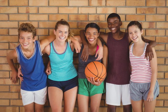 Group of diverse teenagers standing together in a basketball court, smiling and showing camaraderie. Ideal for use in educational materials, sports promotions, youth programs, and advertisements focusing on teamwork and friendship.