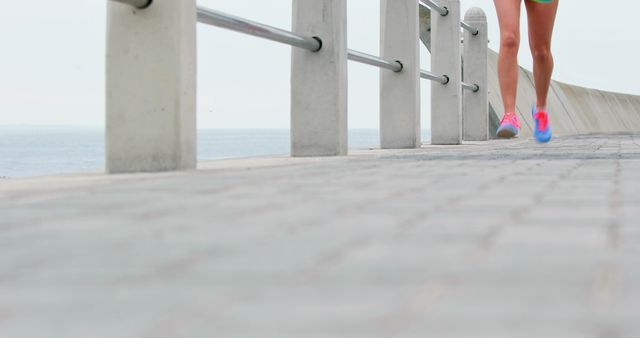 Close-up view of an individual running along a waterfront promenade. Focus on legs in athletic wear, including bright pink shoes. Paved walkway with sea and railing in the background. Ideal for use in fitness, health, and active lifestyle promotions.