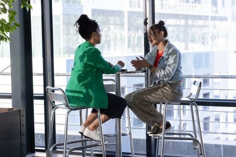 Diverse female coworkers discussing over coffee at modern office lounge with city view
