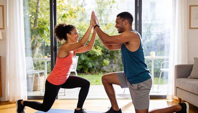 Couple performing lunges and high-fiving during home workout by sunlit patio doors