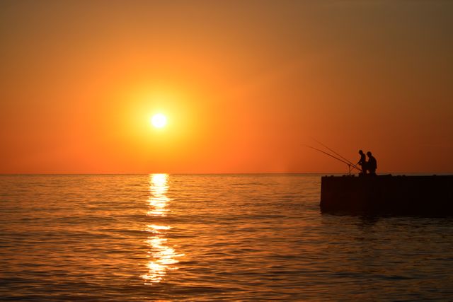Silhouettes of two friends sitting on a pier fishing at sunset, on a calm ocean creating a serene and tranquil atmosphere. Use this image to highlight outdoor leisure activities, friendship moments, or peaceful and scenic nature experiences.