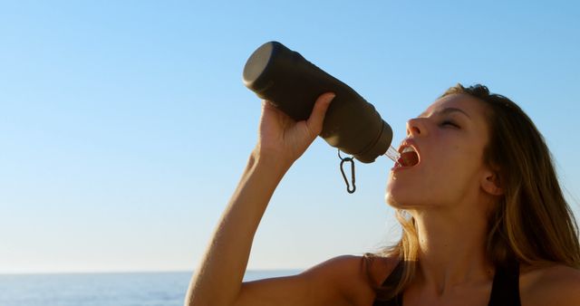 Young woman hydrating from black water bottle with blue sky and sea background. Perfect for promotions related to fitness, hydration, healthy lifestyles, and outdoor activities.