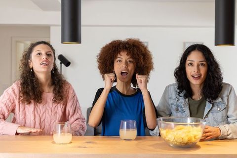 Friends cheering while watching sports at kitchen counter with chips, drinks, casual vibe