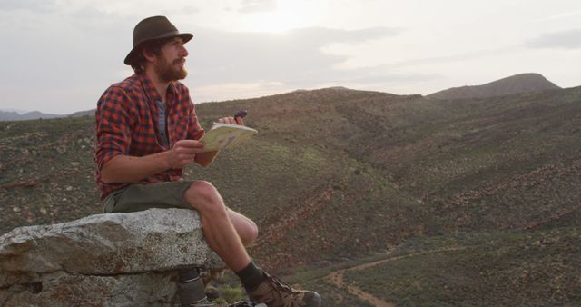 Caucasian male survivalist sitting on mountain peak in wilderness, holding map and compass. exploration, travel and adventure, survivalist in nature.