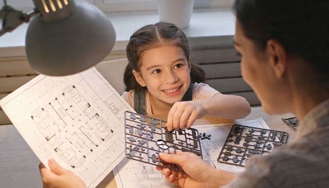 Young girl building miniature architectural model with caregiver studying blueprints