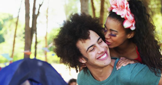 A young African American couple enjoys a playful moment outdoors at a festival, with the woman giving the man a kiss on the cheek. Their joyful expressions and casual attire suggest a relaxed and happy atmosphere at the event.